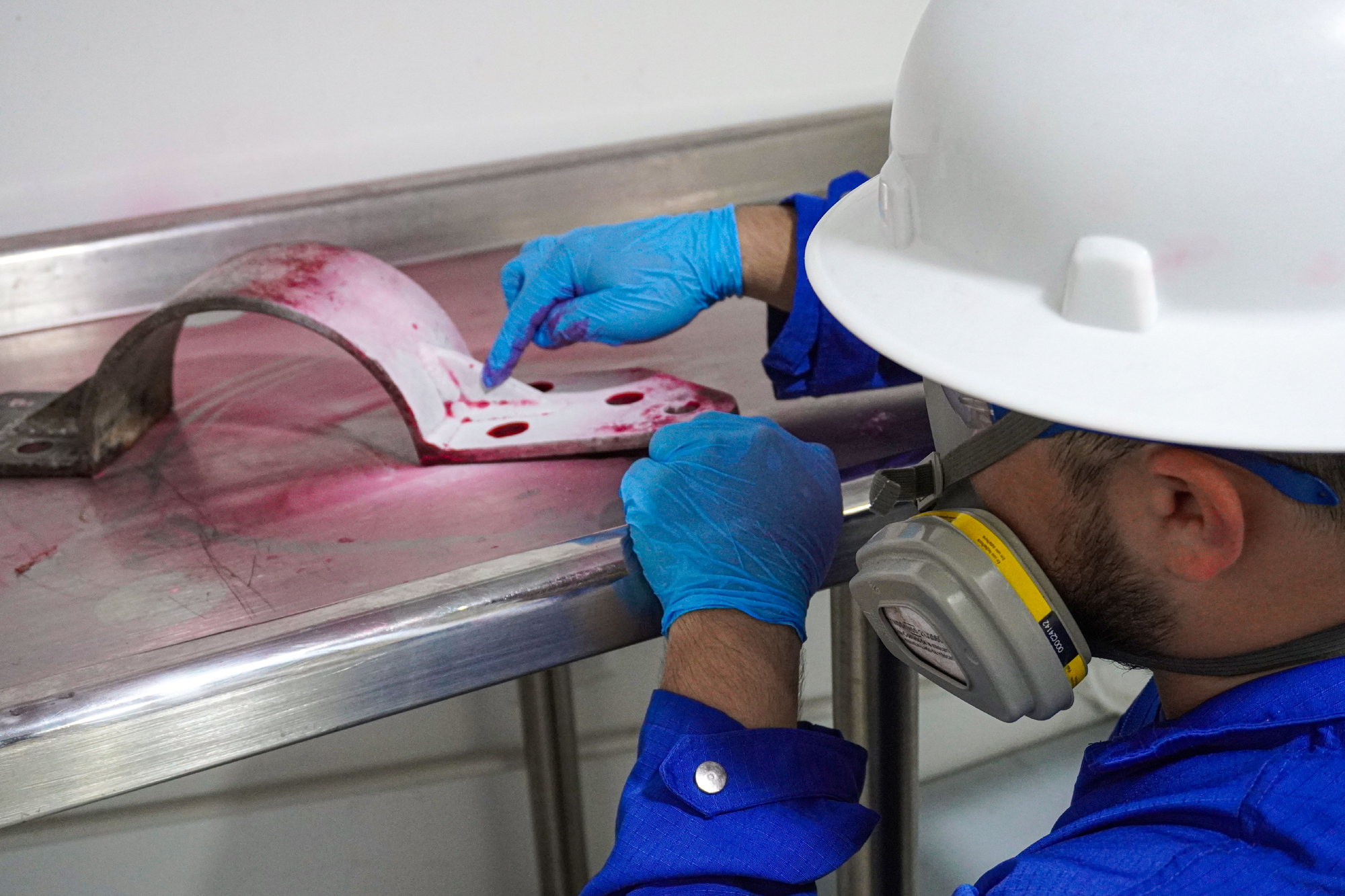 Inspector con casco, gafas y protección respiratoria aplicando líquido penetrante rojo sobre una platina metálica en la mesa de trabajo del laboratorio.