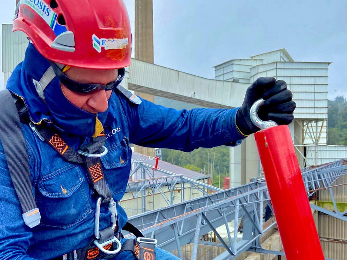 Técnico con arnés y casco ajustando un poste de anclaje rojo sobre la cubierta de una planta industrial.