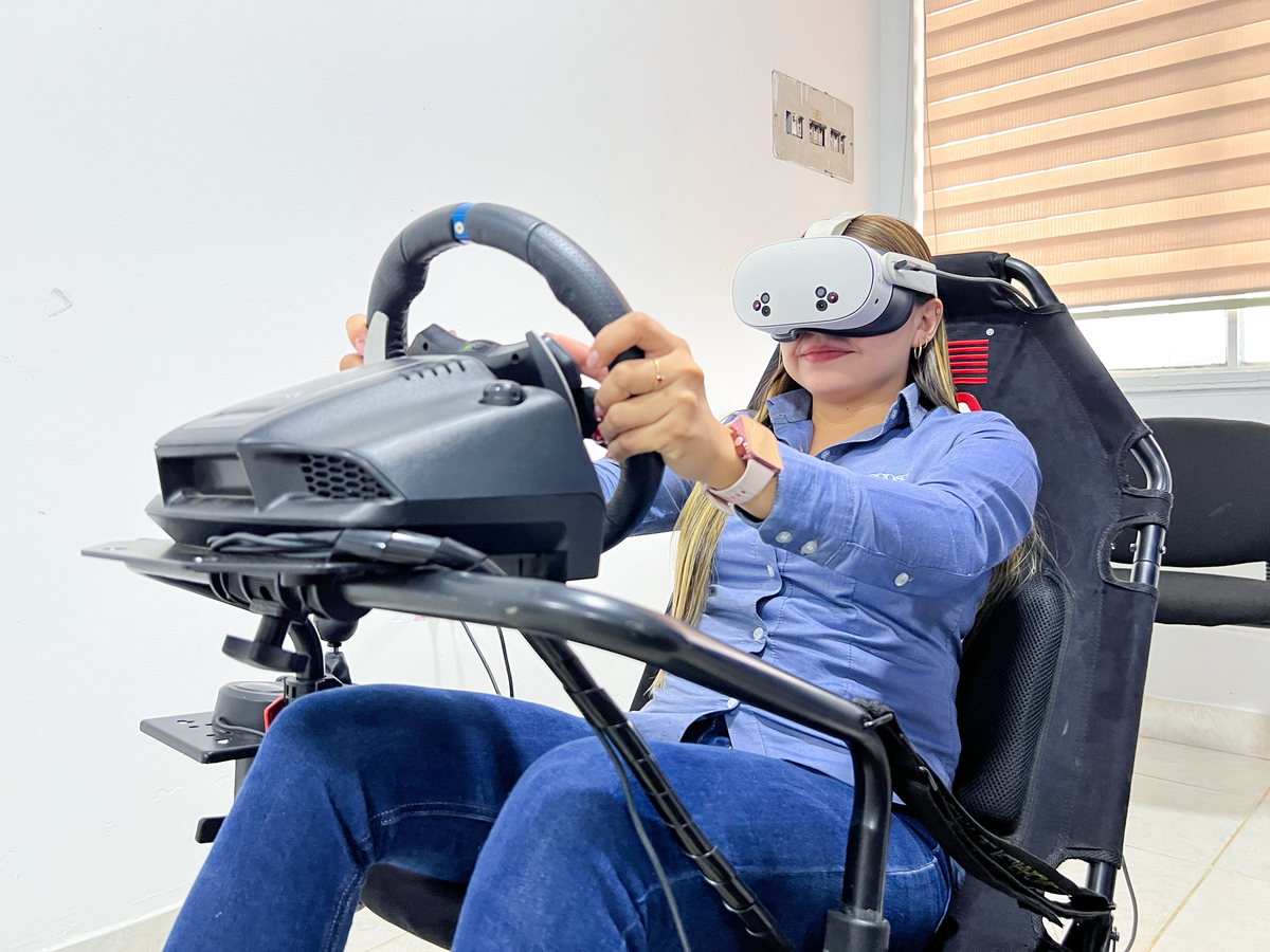 Persona con visor de realidad virtual sujetando el volante en el puesto de simulación de conducción, sentada en la butaca del equipo.
