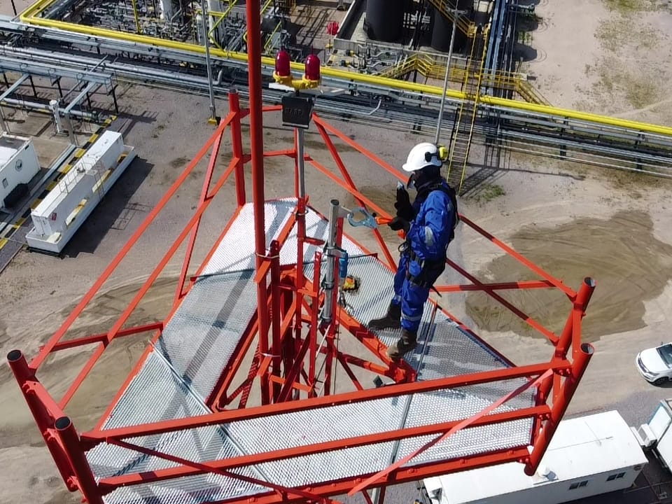 Inspector con casco y arnés de cuerpo entero sobre la plataforma superior de una torre metálica, con la planta industrial abajo.