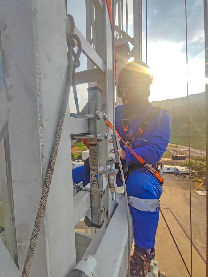 Trabajador con arnés sube por una escalera fija junto al cable de la línea de vida, a contraluz.