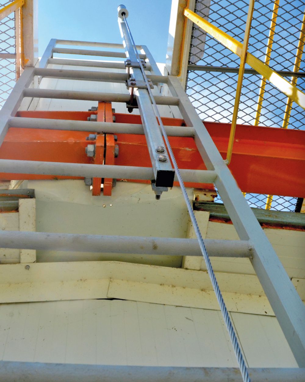 Vista hacia arriba de una escalera fija con el cable de acero de la línea de vida recorriéndola y el mástil galvanizado en la parte superior.