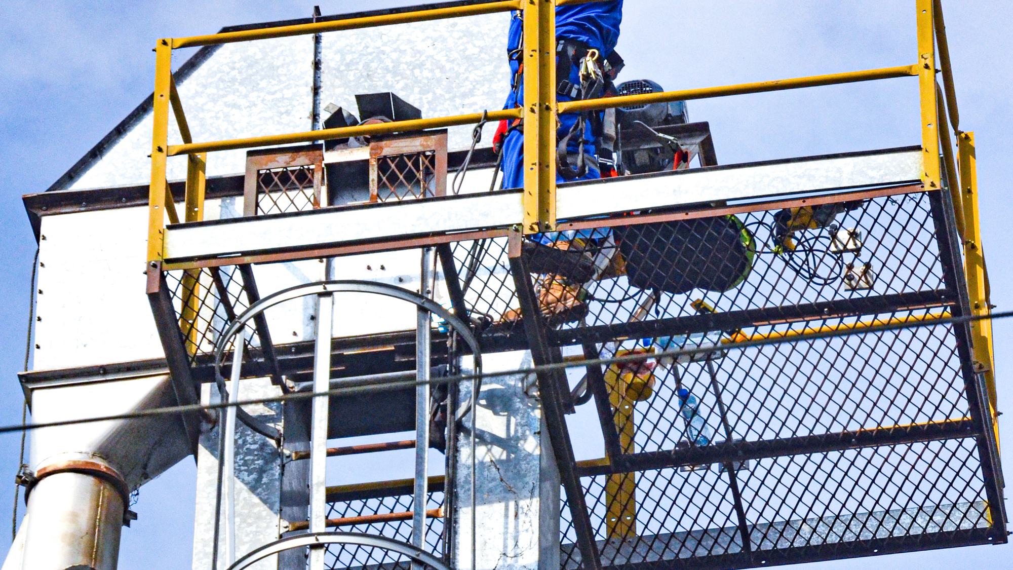 Plataforma superior de una torre metálica, con un trabajador en lo alto de la escalera equipada con línea de vida vertical, contra el cielo azul.