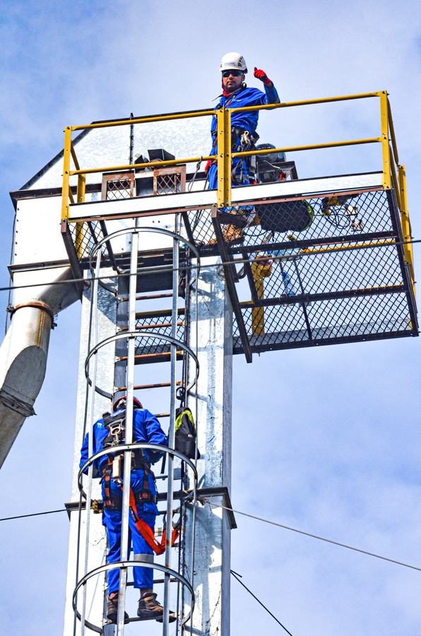 Trabajador sube por la escalera con jaula de una torre metálica hacia la plataforma superior, donde lo espera un compañero.